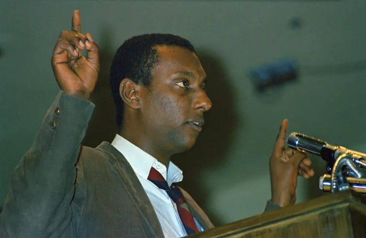 A man speaks at a lectern with fingers pointed up.