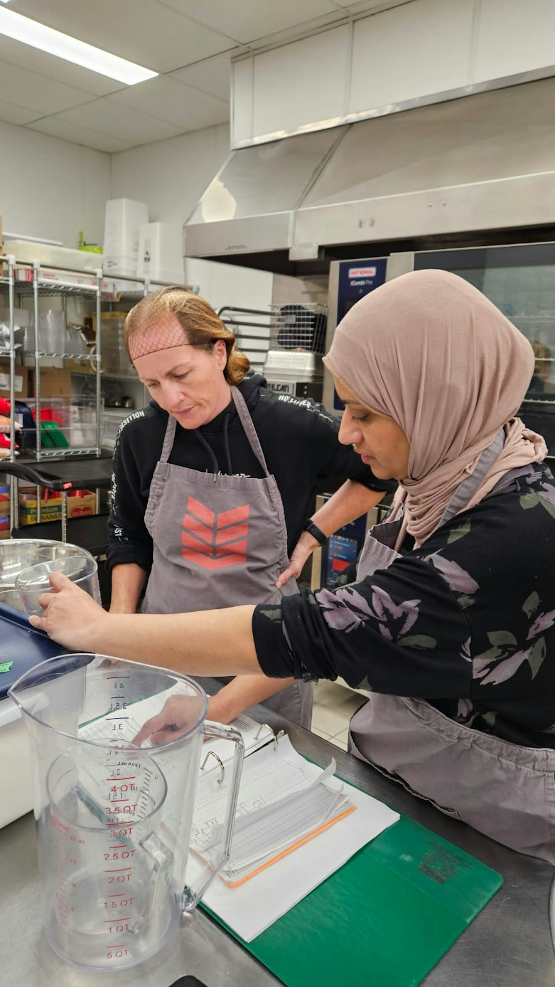 Two adults in a kitchen, one in a hairnet and one in a hijab, scoop food into a container.