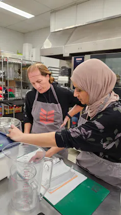 Two adults in a kitchen, one in a hairnet and one in a hijab, scoop food into a container.