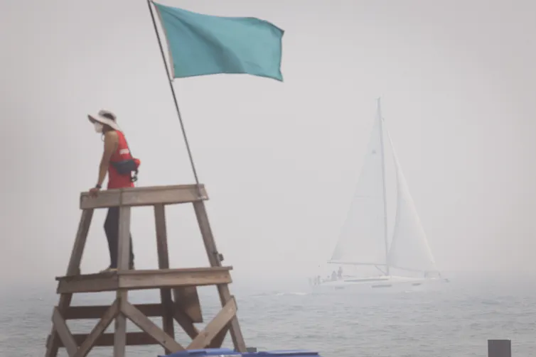 A lifeguard on a stand on a beach. The smoke is so thick, a sailboat is barely visible in the background.