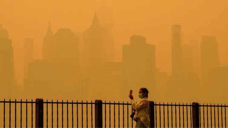 A person walks along the river holding a phone. Buildings that are normally clearly visible disappear into the haze in New York City.