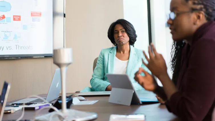 A Black woman in a suit sits at the head of a conference table, watching another woman speak