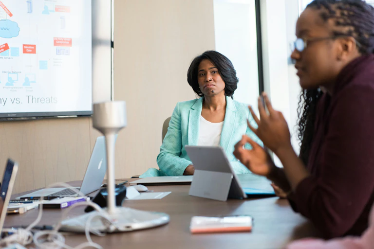 A Black woman in a suit sits at the head of a conference table, watching another woman speak