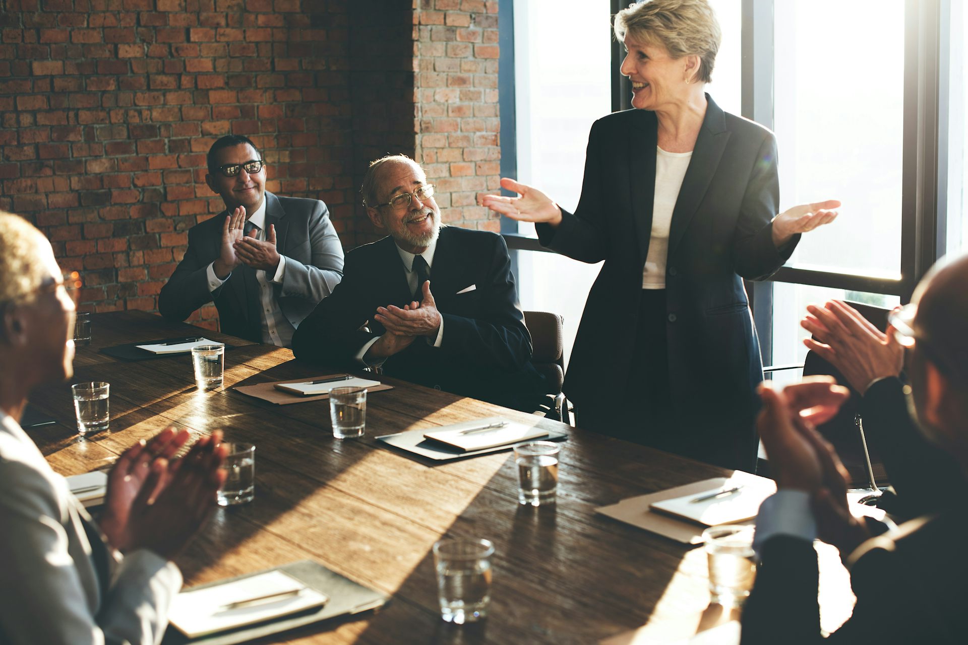 A woman in a suit stands while speaking to a group of people seated around a conference table