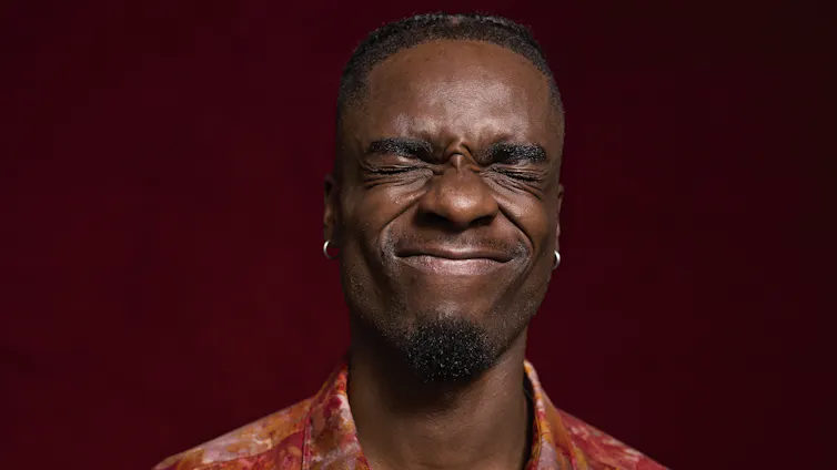 A young person with eyes closed makes a face in front of a maroon background