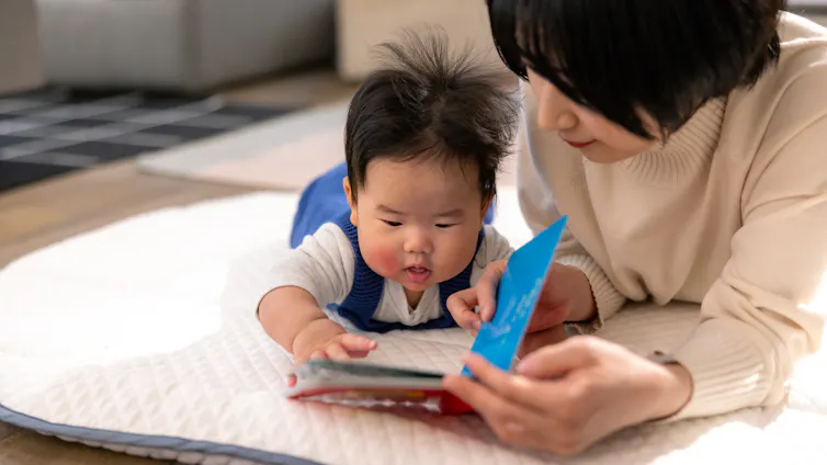 Baby boy and her mother reading a book in living room.