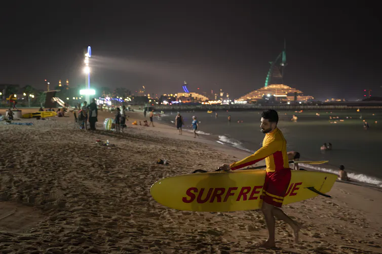 lifeguard with surfboard walking up beach at nighttime, floodlights behind.