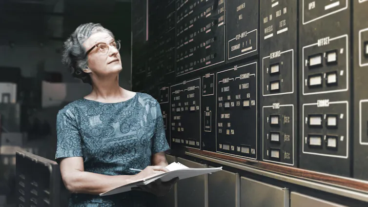 A woman with grey hair, wearing a blue dress and taking notes in front of a black board.