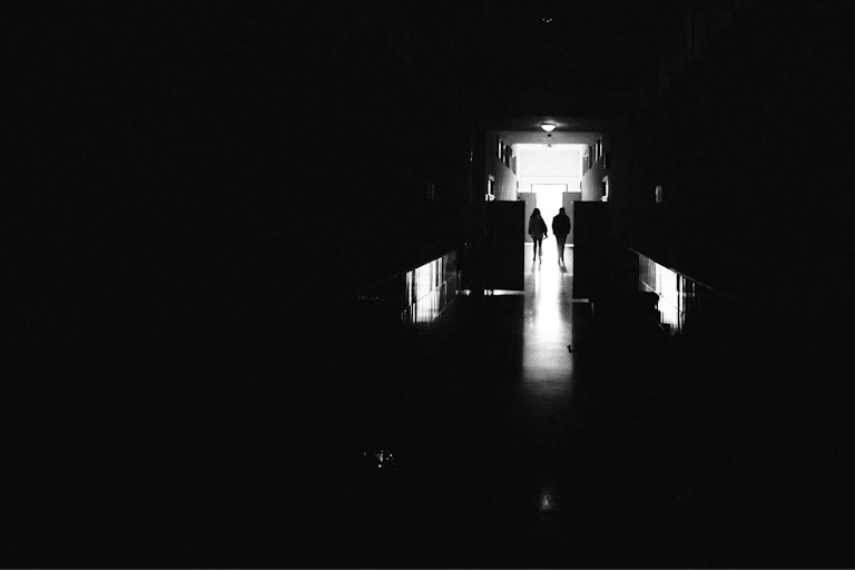 Two people walk down a dark hallway lit by only one light