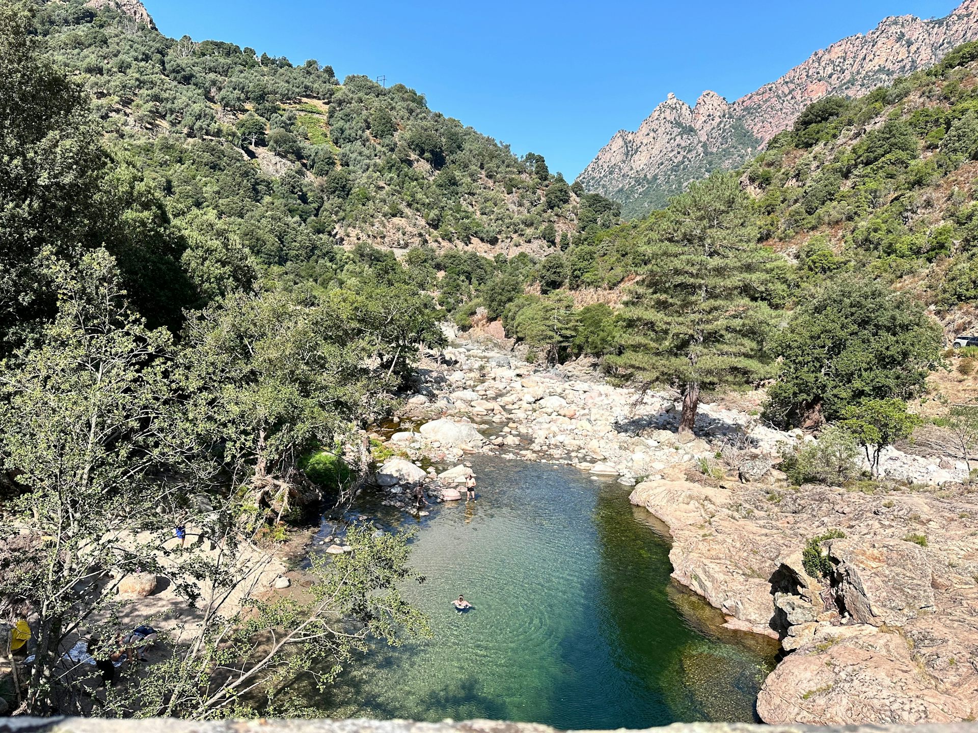 Un paysage montagneux méditerranéen avec au premier plan un bassin naturel dans lequel se baignent deux personnes.