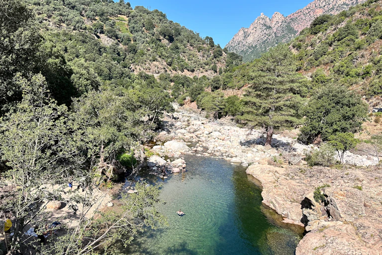 Un paysage montagneux méditerranéen avec au premier plan un bassin naturel dans lequel se baignent deux personnes.