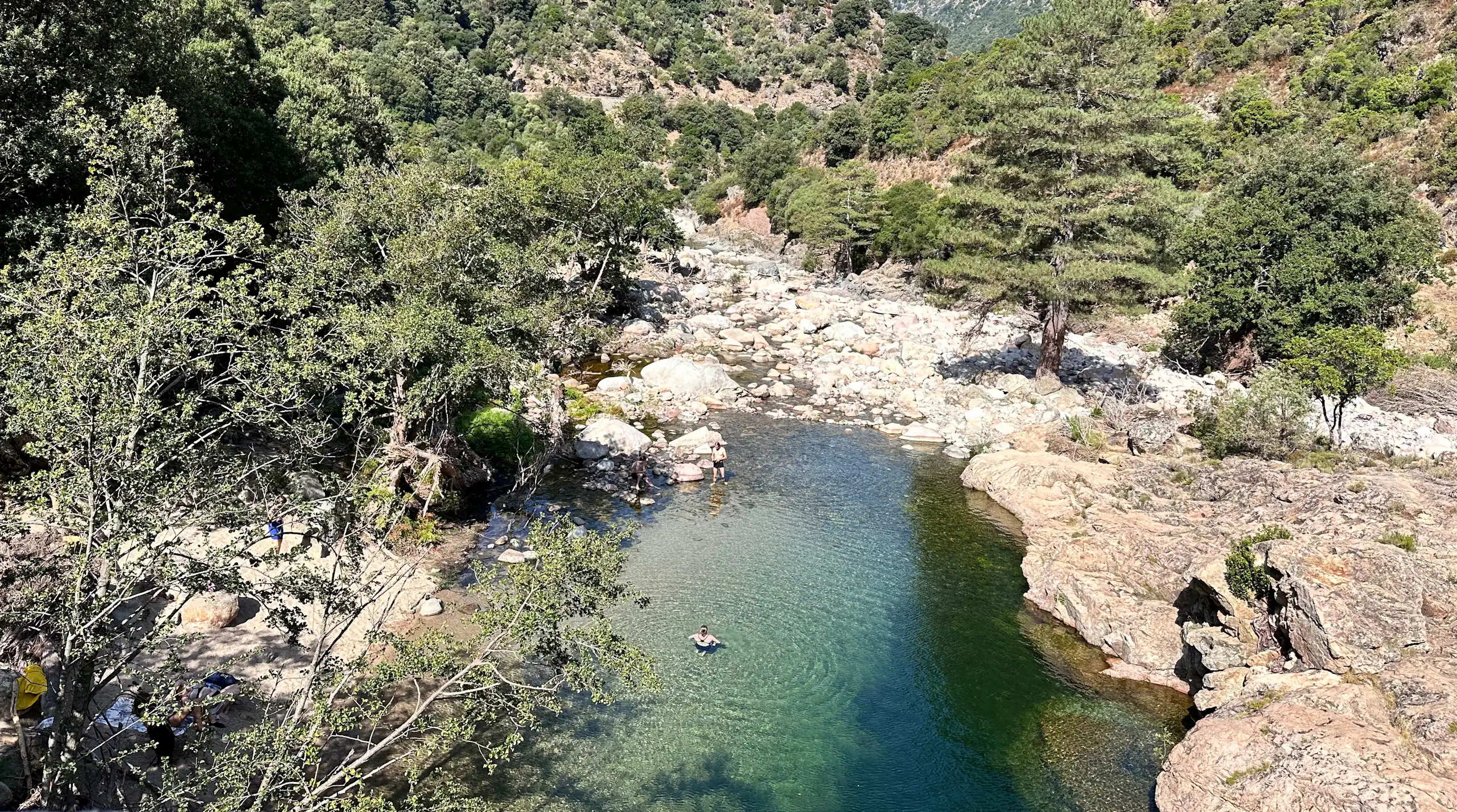 Un paysage montagneux méditerranéen avec au premier plan un bassin naturel dans lequel se baignent deux personnes.