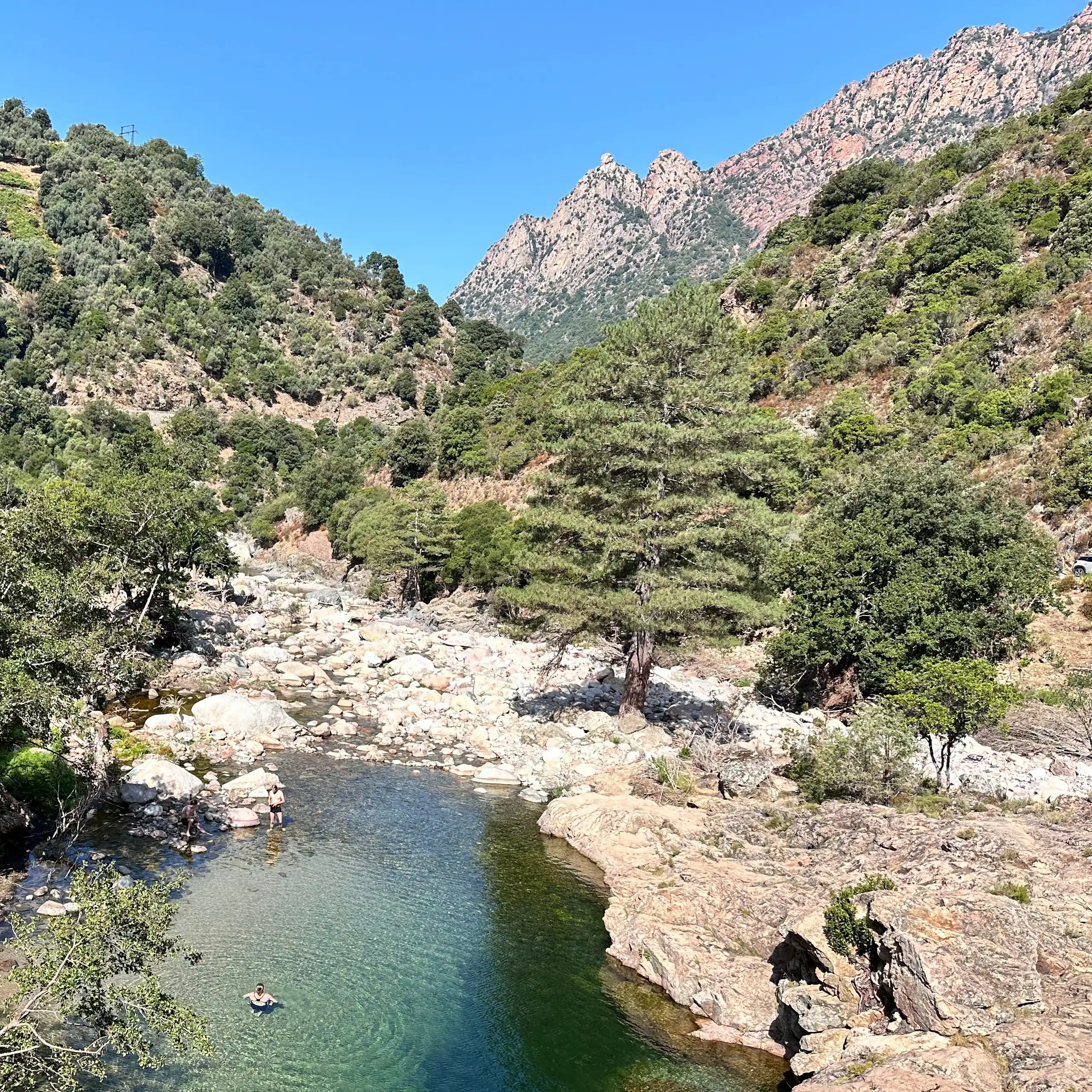 Un paysage montagneux méditerranéen avec au premier plan un bassin naturel dans lequel se baignent deux personnes.