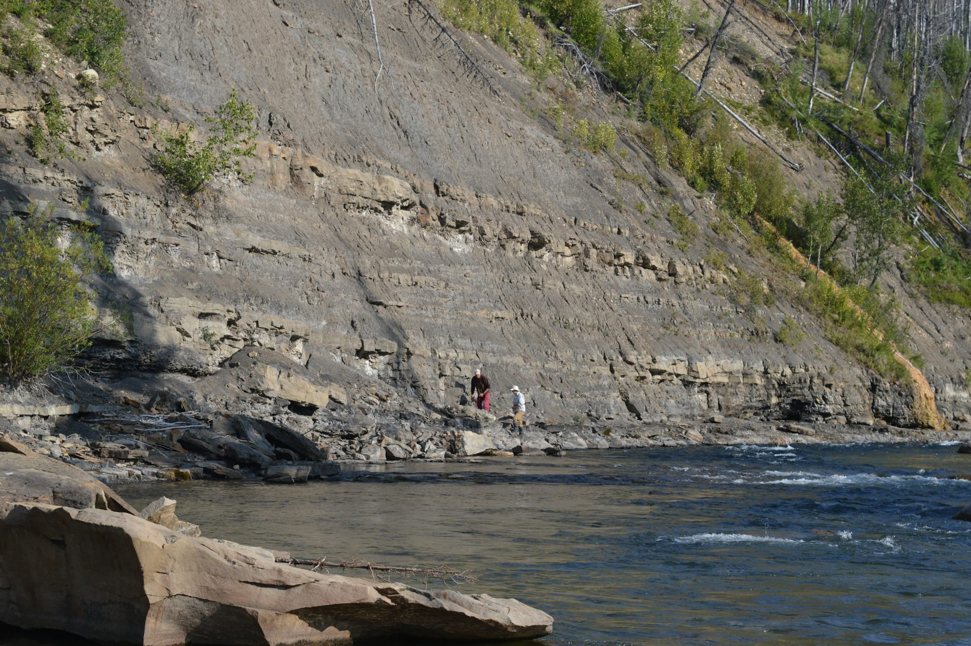 A steep rock face against a body of water, with two distant men walking beneath.