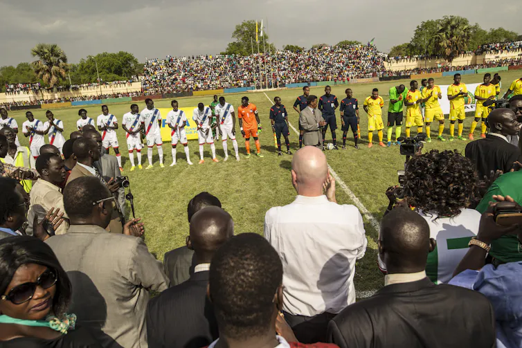 Un homme européen chauve photographié de dos au milieu d'une petite foule qui regarde deux équipes sportives africaines et des officiels s'aligner sur un terrain de sport.