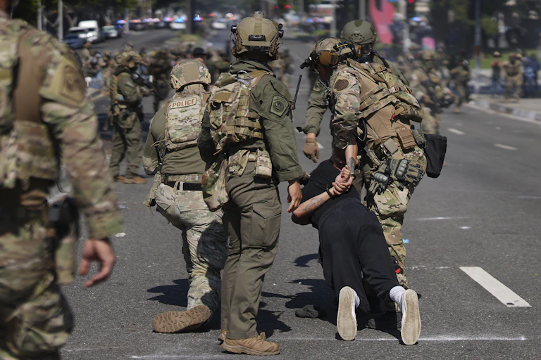 Officers in camouflage gear and helmets detain a man, his hands held behind his back, as rows of uniformed personnel line the road behind them.