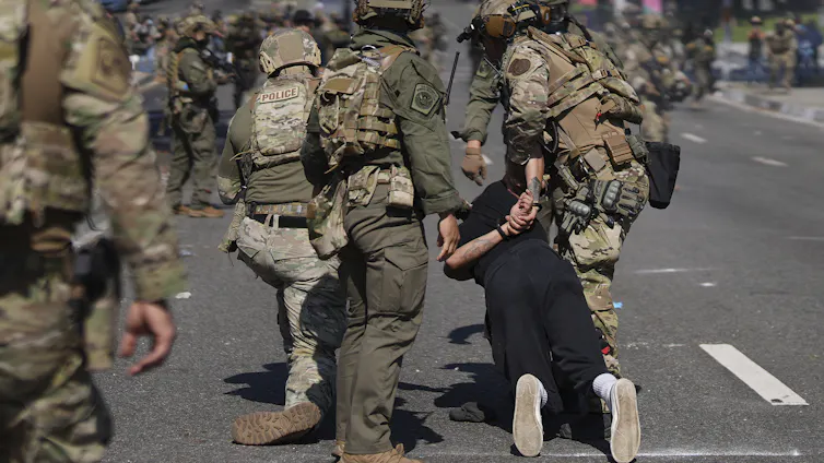 Officers in camouflage gear and helmets detain a man, his hands held behind his back, as rows of uniformed personnel line the road behind them. Human Rights