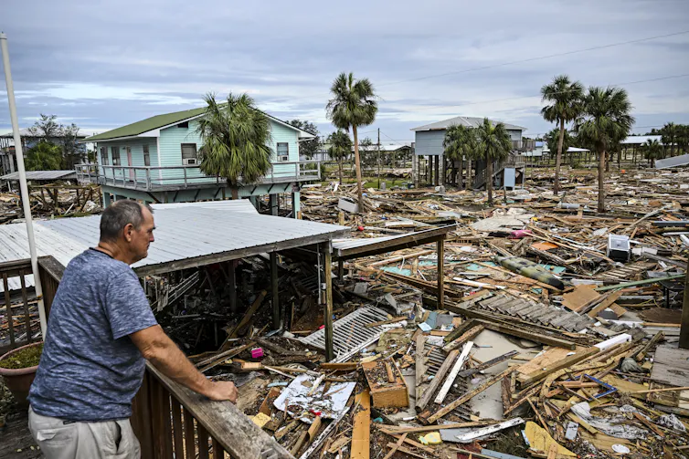 A man stands at a railing after a hurricane, looking out at a sea of scattered wood and debris that was once part of buildings. A few houses on stilts are still standing in the background.
