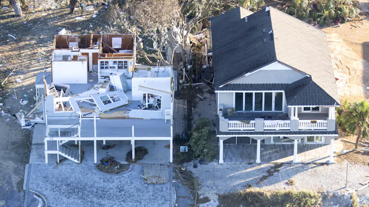 Two homes side by side, one mostly untouched, the other largely destroyed, with walls torn out and the roof gone.