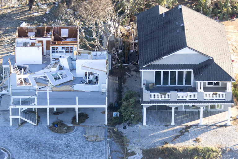 Two homes side by side, one mostly untouched, the other largely destroyed, with walls torn out and the roof gone.