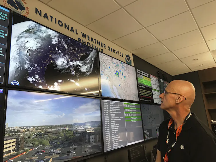 A scientist looking at six TV screens showing lists of data, maps of storms and radar