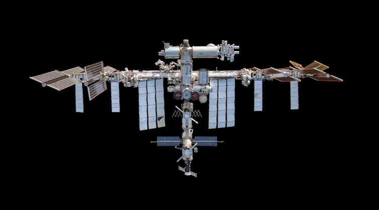 The International Space Station, which is a metal structure with solar panels coming off it, floating above Earth