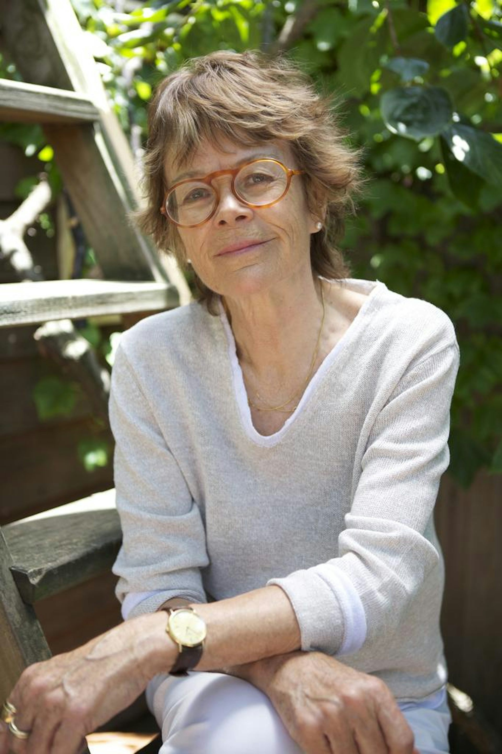 Hilary McPhee, a woman with short hair and glasses, sitting on a ladder in front of greenery.