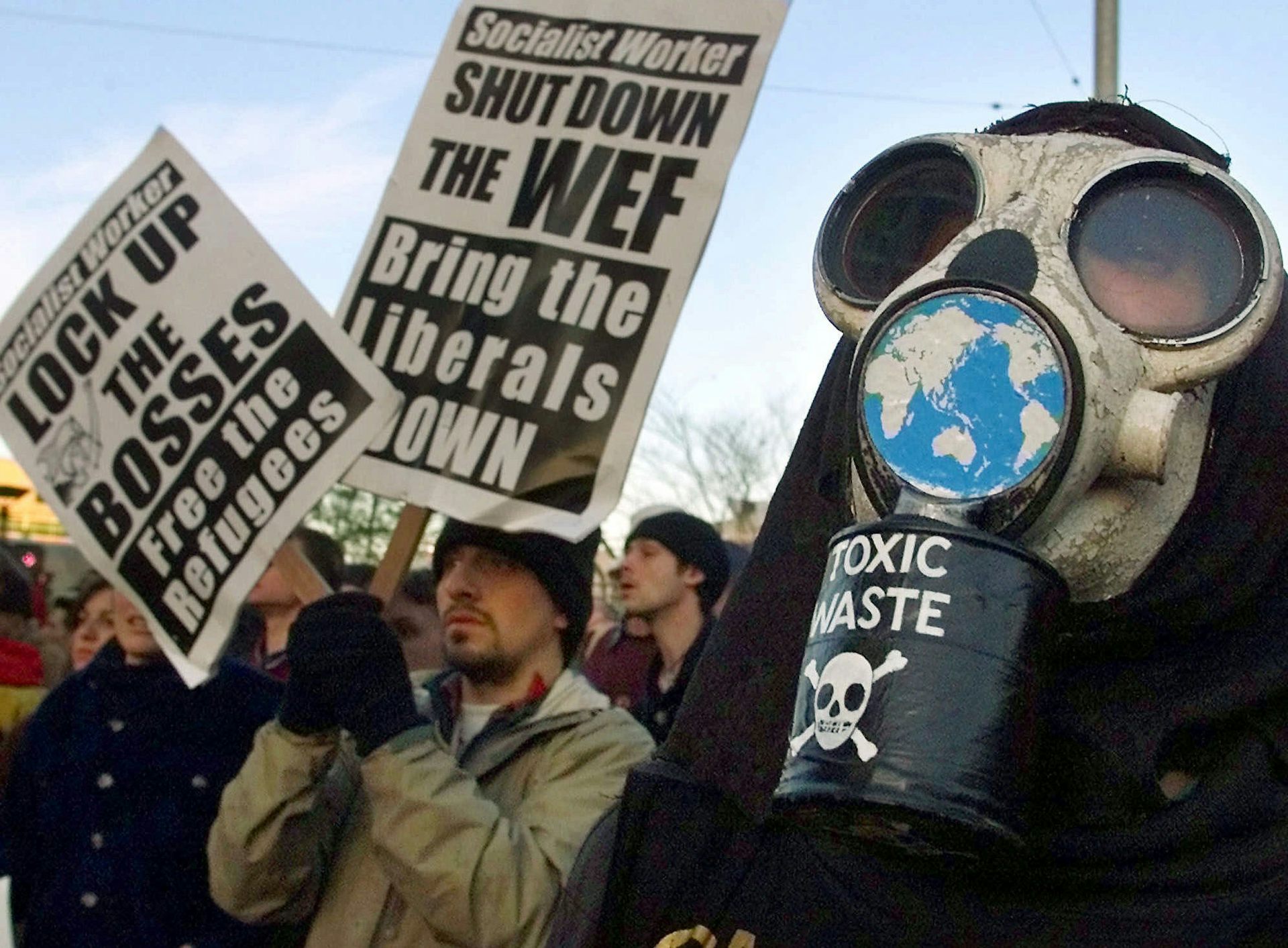 A person wearing a gas mask and another holding two signs reading 'shut down the WEF' and 'Lock up the bosses'