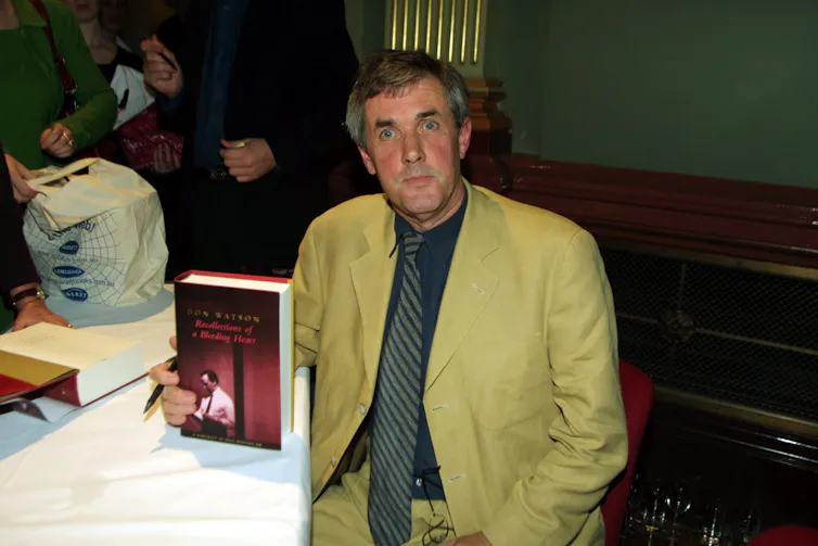 Don Watson, author of Recollections of a Bleeding Heart A Portrait of Paul Keating PM, at the book's launch, holding a copy. He wears a light greenish suit with a black shirt and tie.