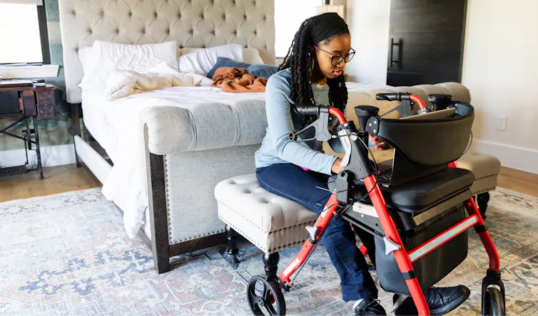 A young woman uses her laptop while sitting on a walker in her bedroom.