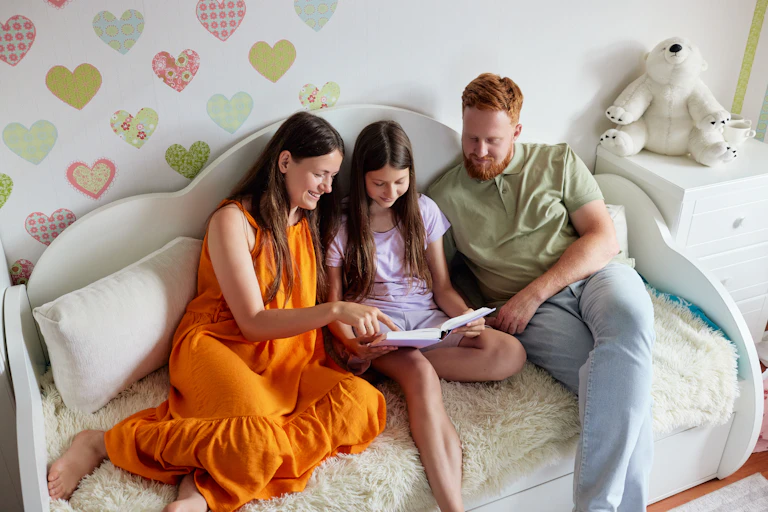 A mother, father and their daughter sit on a white sofa, reading a book together.