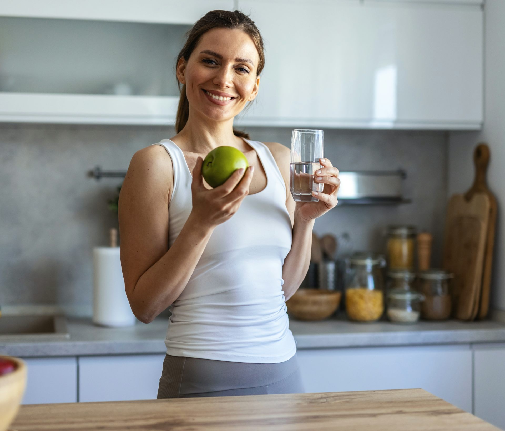 Une jeune femme sourit en tenant, d'une main, un verre d'eau et, de l'autre, une pomme verte.