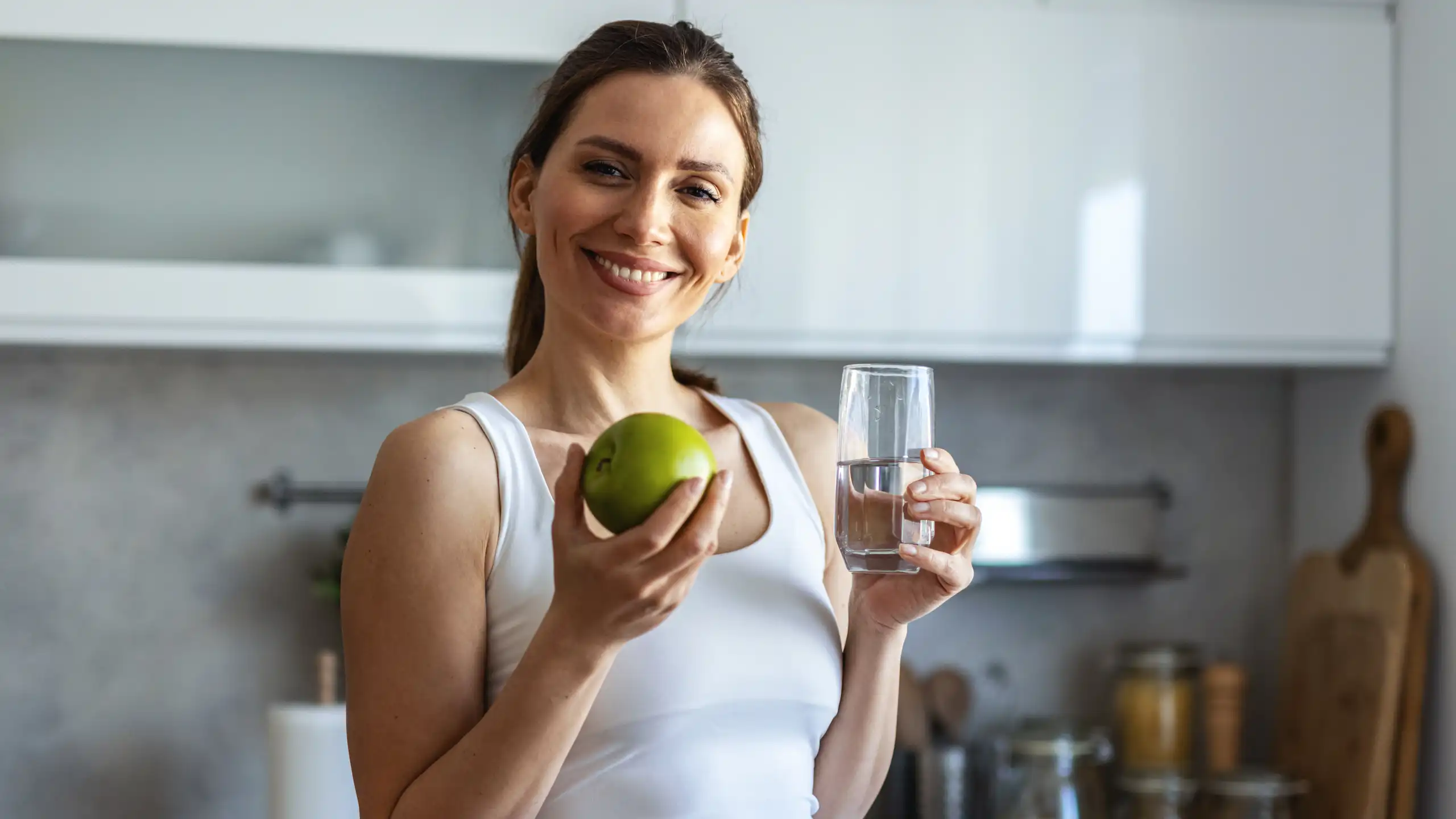 Une jeune femme sourit en tenant, d'une main, un verre d'eau et, de l'autre, une pomme verte.