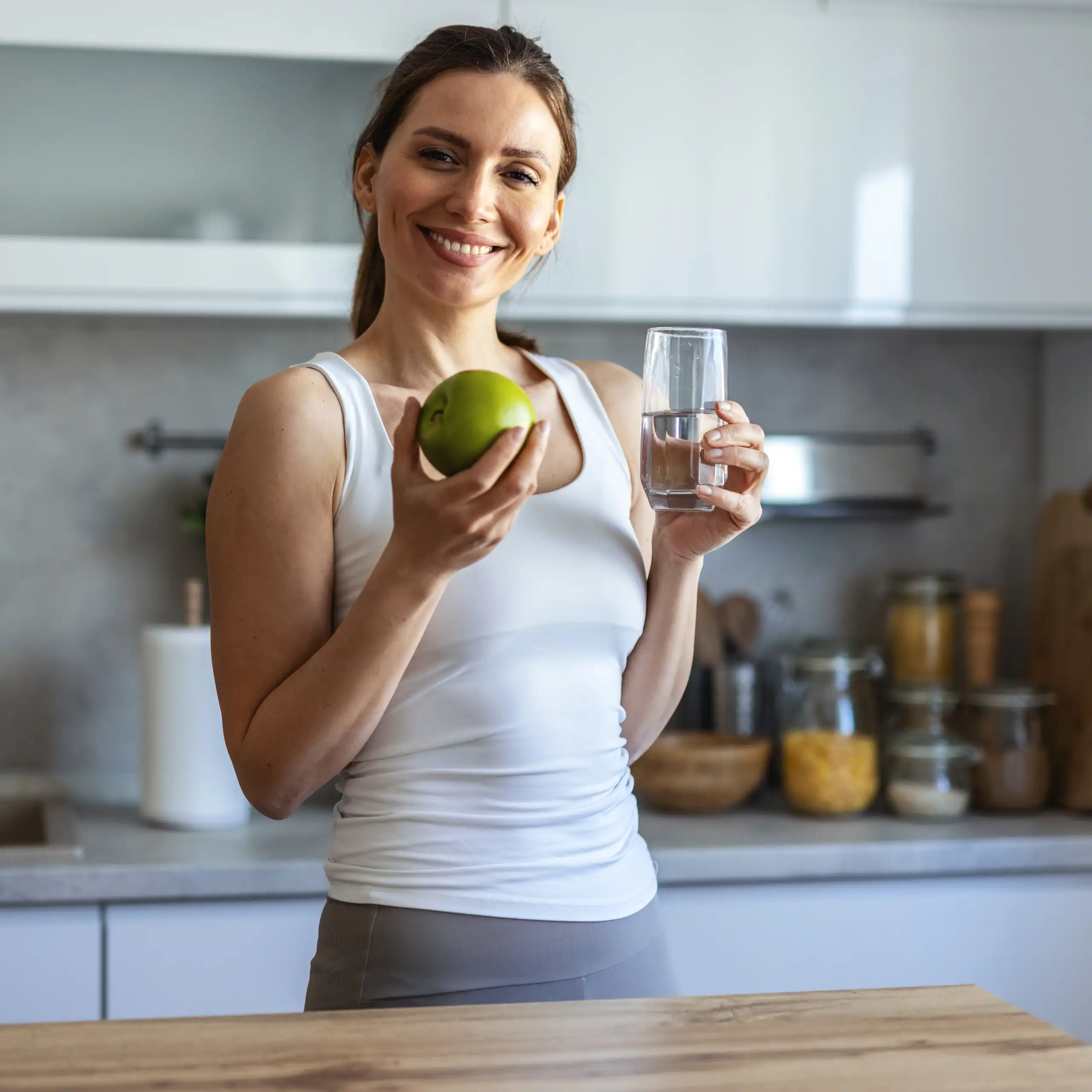 Une jeune femme sourit en tenant, d'une main, un verre d'eau et, de l'autre, une pomme verte.