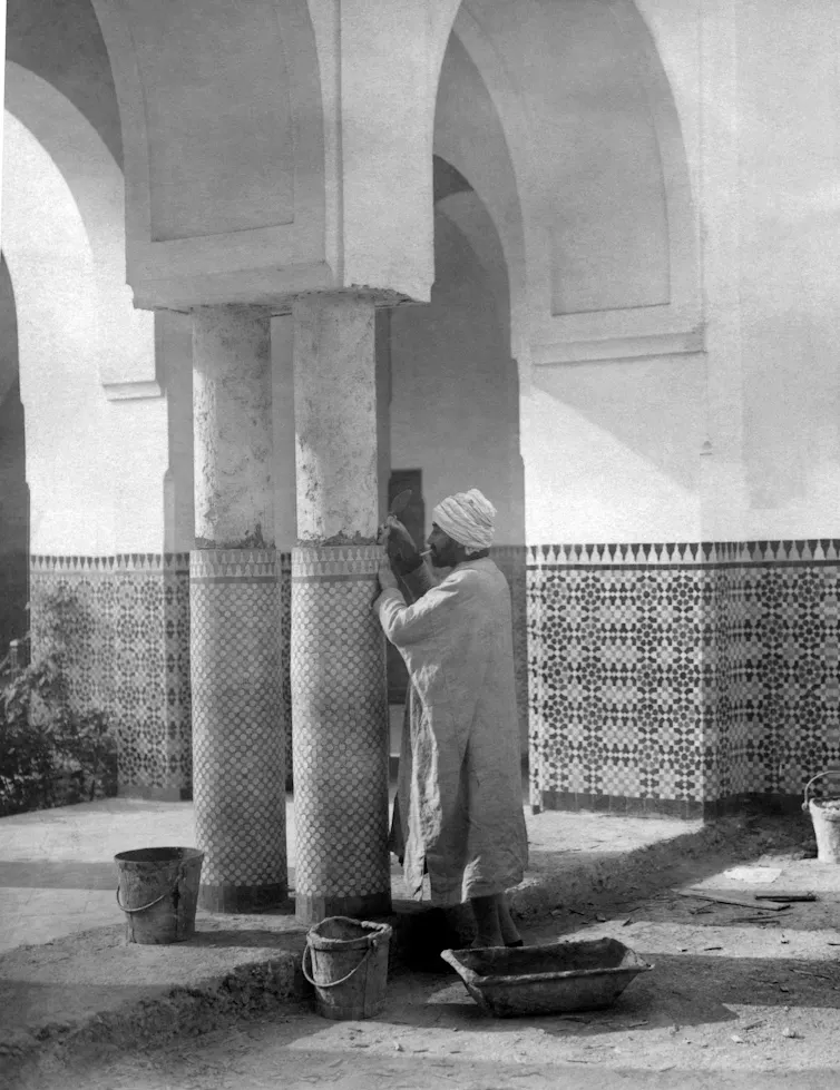 A man wearing a head covering and a long shirt looks intently at a pillar beneath an archway, with several buckets nearby.