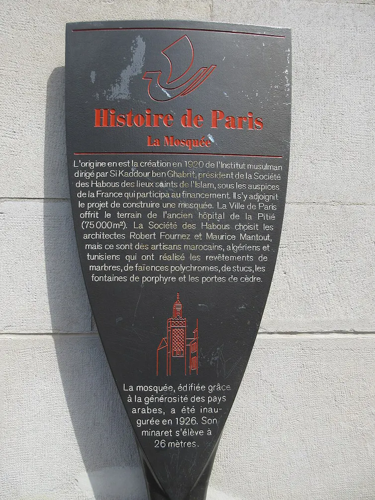 A black, shield-shaped plaque mounted on a light-colored stone wall, with red and white French text.