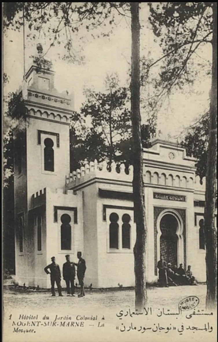 A historic black-and-white photograph of a small mosque with a minaret, arched windows and a large arched entrance, surrounded by trees.