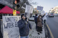 People stand on a road with signs saying 'We stand together for fair water access' and 'United for water equity'