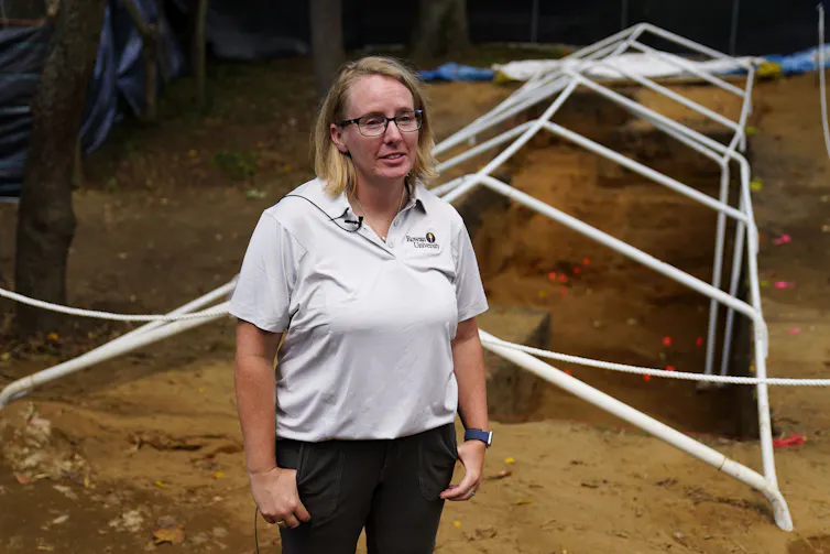 A woman wearing a light-colored polo shirt and black pants stands in an area surrounded by dirt