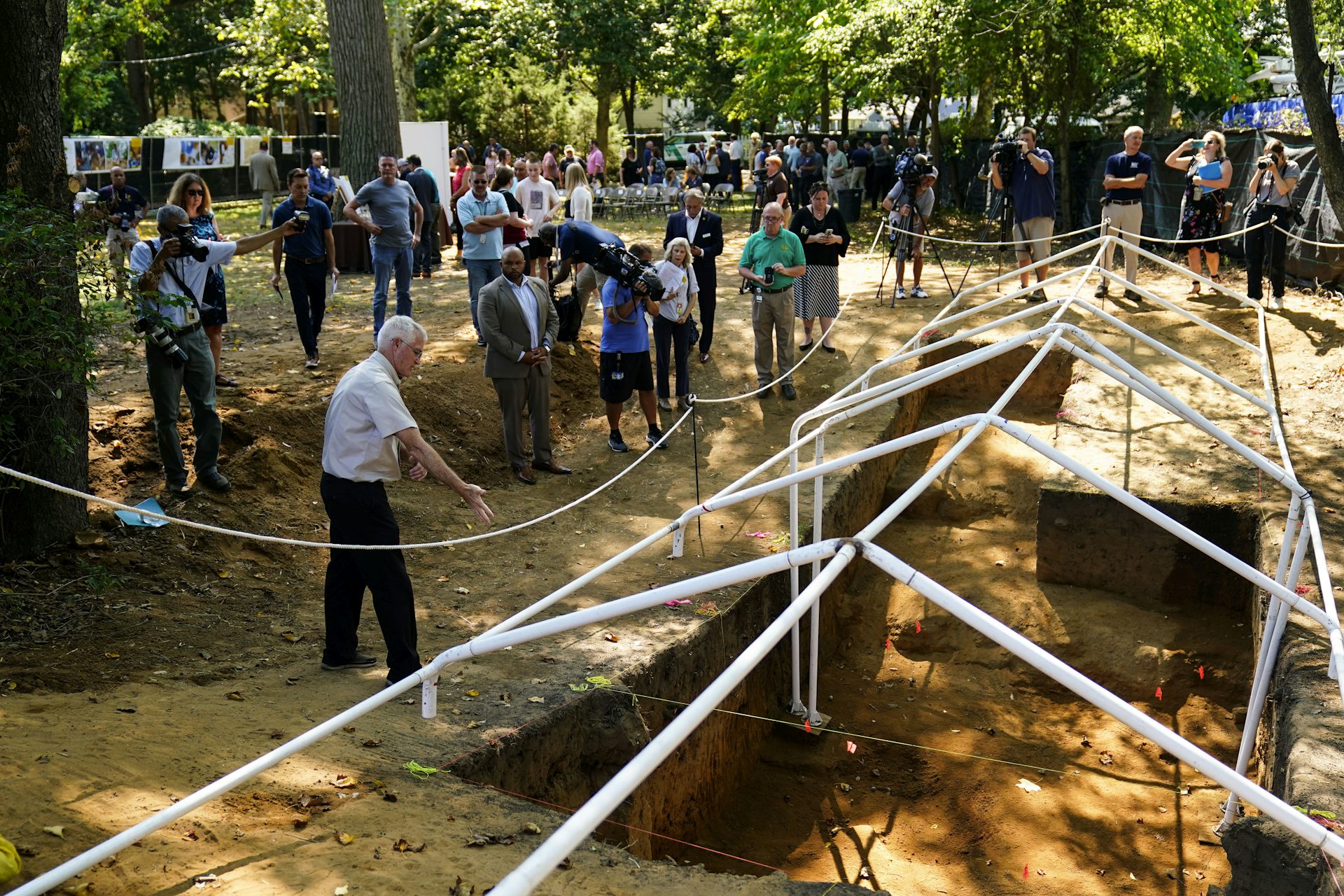 A group of people, several holding cameras, stand around a large hole dug in the ground