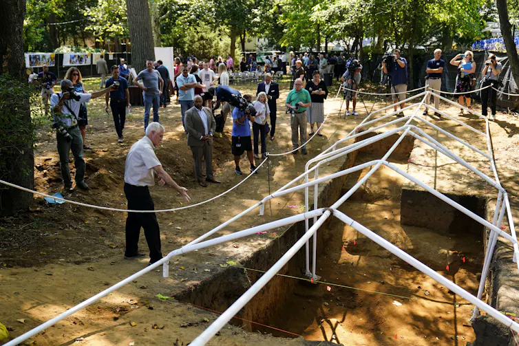 A group of people, several holding cameras, stand around a large hole dug in the ground