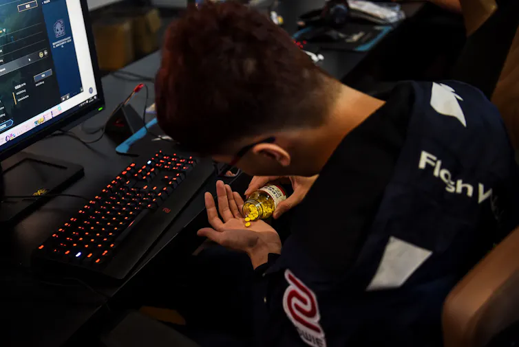 Young man wearing a black sweatshirt pours yellow pills into his open palm while seated in front of a computer monitor and keyboard. Professional Gaming.