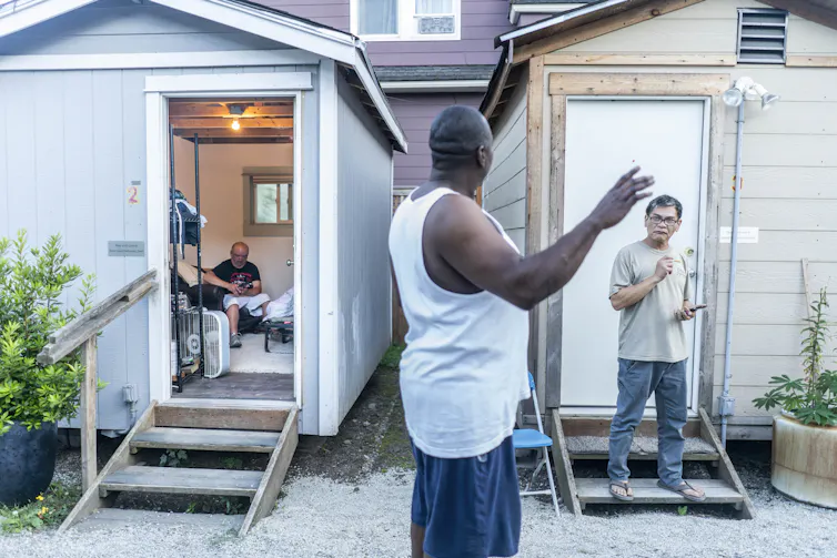A man speaks to another man standing on the steps of a small house.