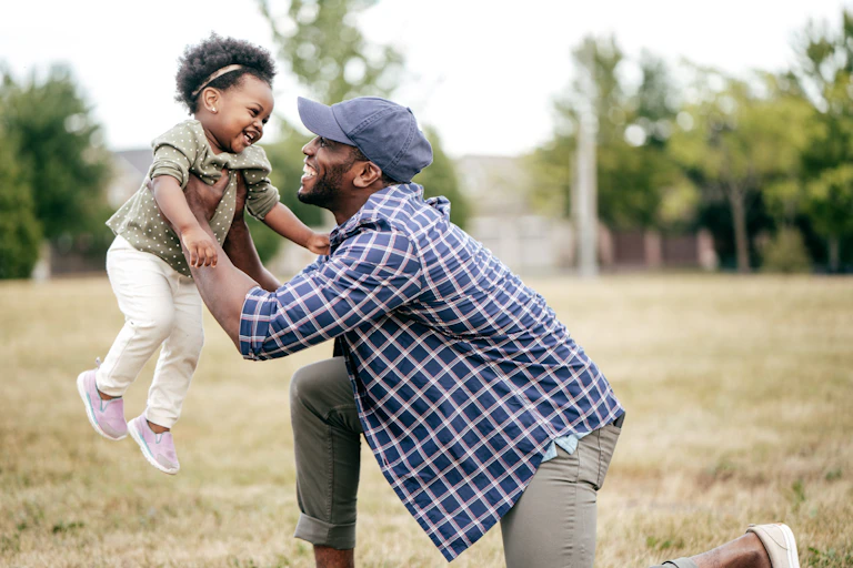 A father kneeling in a field smiling broadly holding his daughter up in the air while she laughs