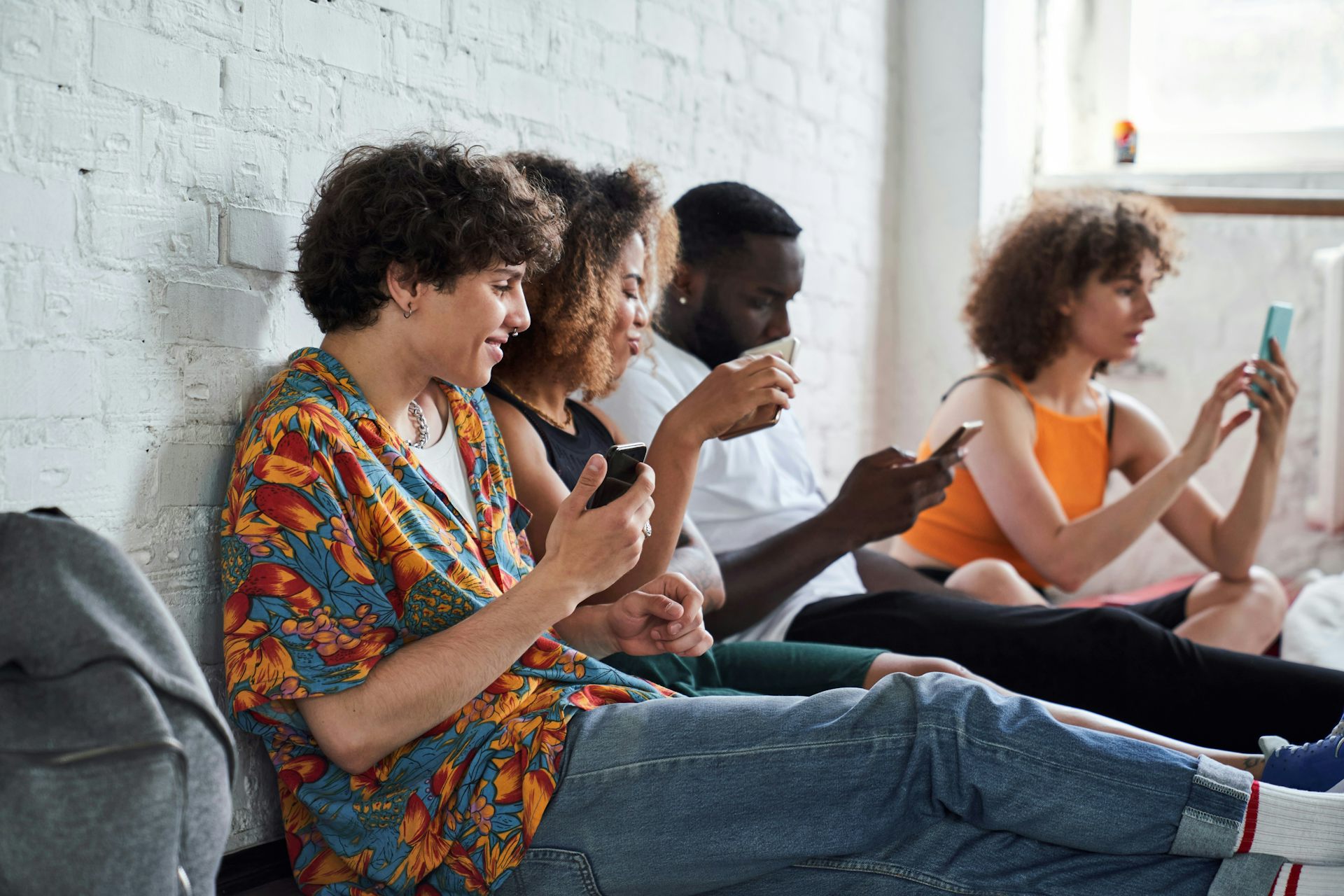 Youth sitting with their backs against a wall looking at phones.