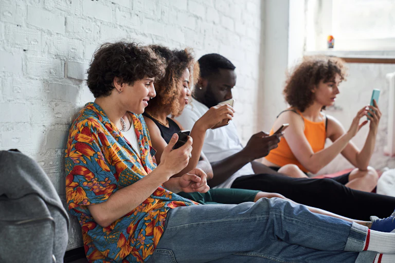 Youth sitting with their backs against a wall looking at phones.
