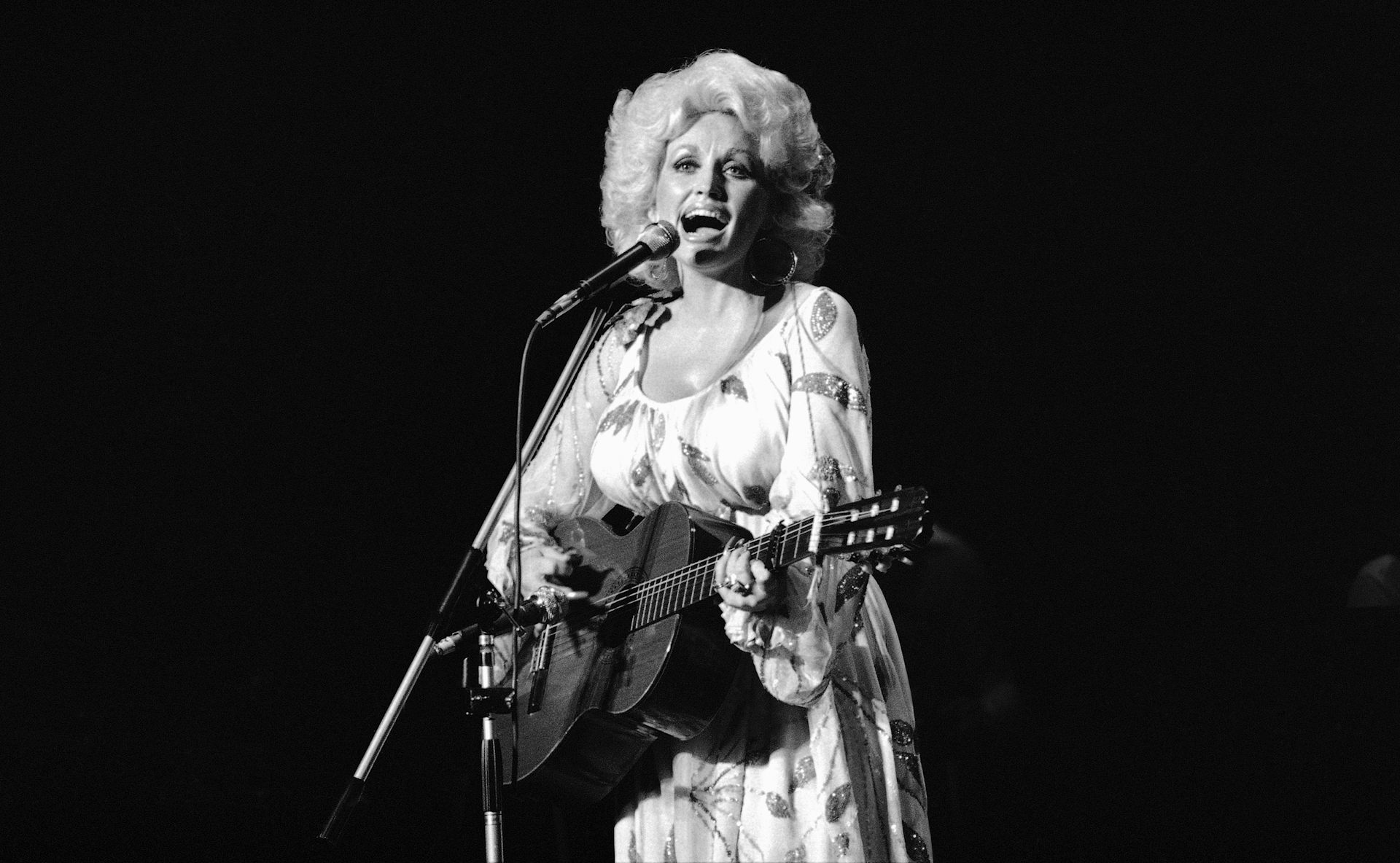 Black and white photo of a blonde woman in a dress playing a guitar and singing into a microphone