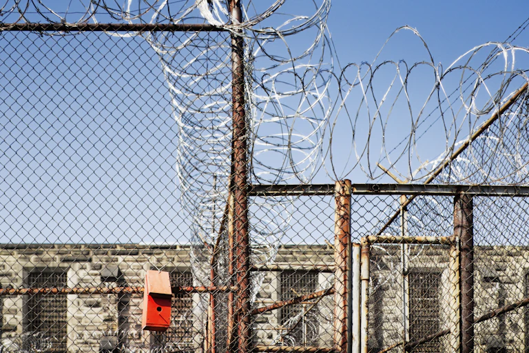 A small red birdhouse hangs on a chain-link fence topped with razor wire outside a stone building.