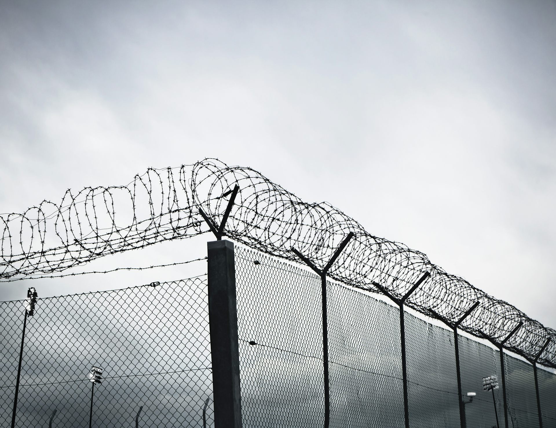 A grayscale view of a tall chain-link prison fence topped with rows of barbed wire
