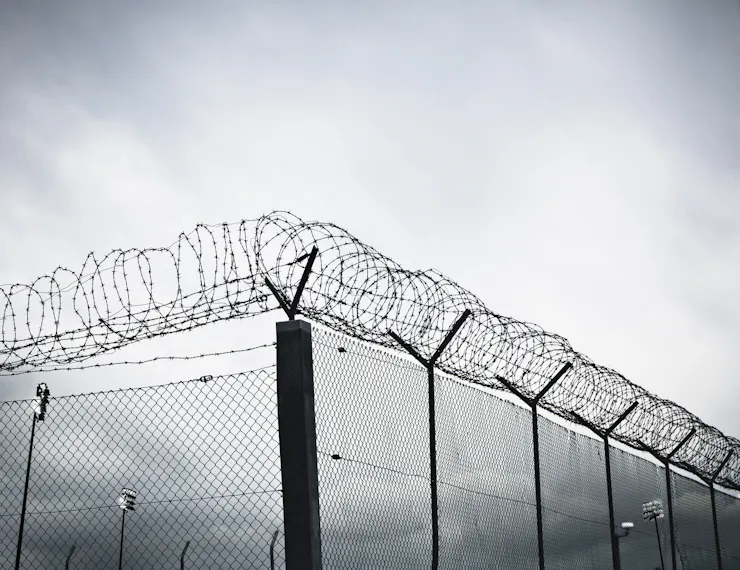 A grayscale view of a tall chain-link prison fence topped with rows of barbed wire